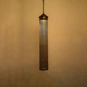 May include: A cylindrical pendant light fixture with a brass finish. The light has a patterned, openwork design and hangs from a chain. The fixture is suspended against a warm-toned wall.