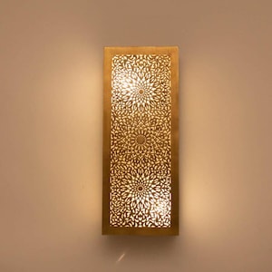 Puede incluir: Aplique de pared rectangular dorado con un intrincado diseño floral. El aplique está iluminado desde el interior, proyectando un brillo cálido. El diseño presenta formas recortadas que crean un efecto de luz decorativo.