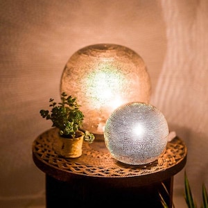 Op de afbeelding: Een decoratieve tafellamp met een bolvormig ontwerp. De lamp heeft een getextureerde, metallic afwerking en straalt een warme gloed uit. Een kleinere, vergelijkbare lamp staat ernaast. Een kleine plant in een pot staat ook op de tafel.