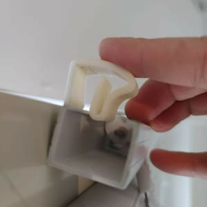 Puede incluir: Primer plano de un componente de persiana de plástico blanco, sostenido por una mano. El componente tiene forma curva y está unido a un mecanismo de persiana gris más grande. La imagen está bien iluminada, mostrando los detalles del plástico.
