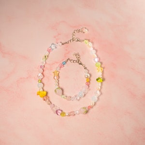 May include: A colorful beaded necklace and bracelet set with a yellow duck charm and a pink strawberry charm. The necklace and bracelet are made of glass beads in various colors, including pink, blue, green, and white.