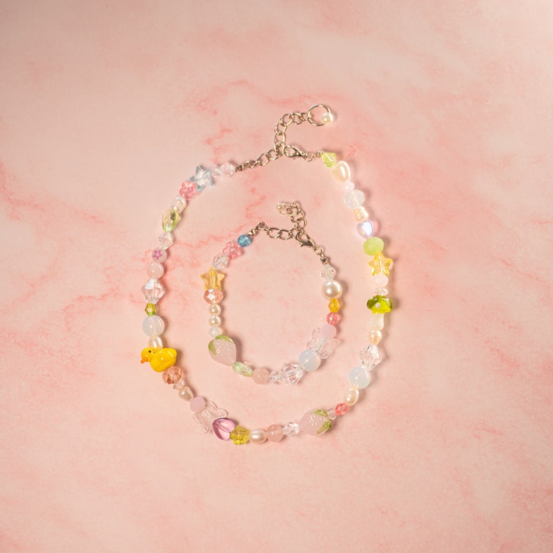 May include: A colorful beaded necklace and bracelet set with a yellow duck charm and a pink strawberry charm. The necklace and bracelet are made of glass beads in various colors, including pink, blue, green, and white.