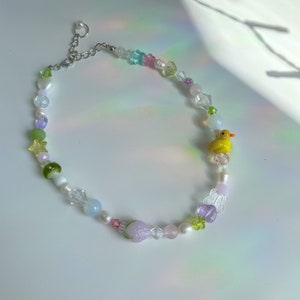 May include: A colorful beaded necklace with a yellow rubber duck charm, a pink strawberry charm, and a clear butterfly charm. The necklace is made with various beads, including clear, white, pink, green, and blue.