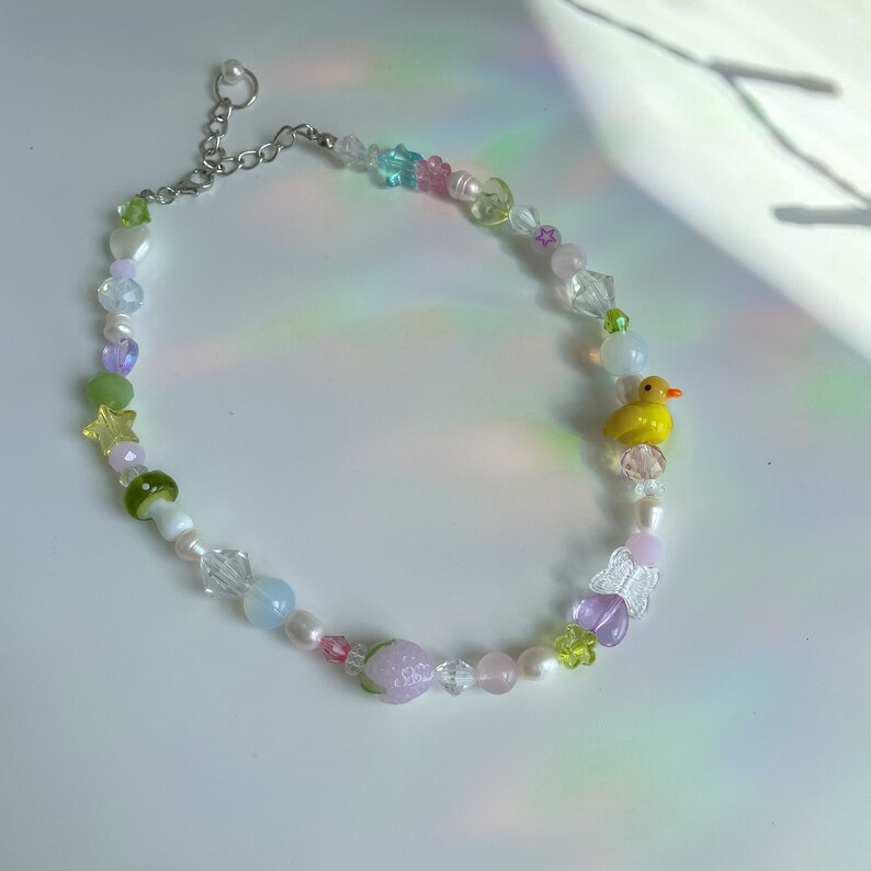 May include: A colorful beaded necklace with a yellow rubber duck charm, a pink strawberry charm, and a clear butterfly charm. The necklace is made with various beads, including clear, white, pink, green, and blue.