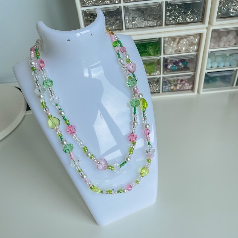 Puede incluir: Un collar de dos hilos con cuentas de color rosa, verde y blanco. El collar est&aacute; en un expositor blanco.