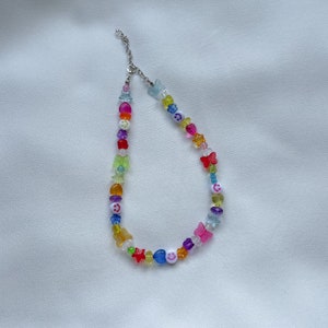 May include: A colourful beaded necklace featuring a range of charms, including butterflies, stars, and smiley faces. The necklace is made from clear plastic beads and has a silver clasp.