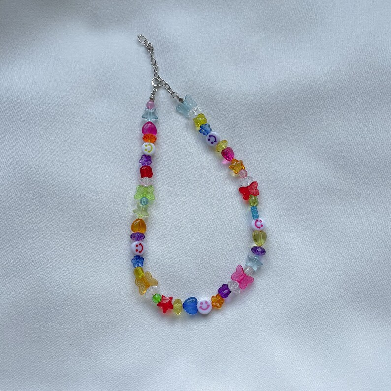 May include: A colourful beaded necklace featuring a range of charms, including butterflies, stars, and smiley faces. The necklace is made from clear plastic beads and has a silver clasp.