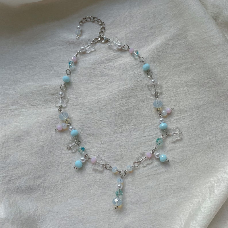 Pode incluir: Um delicado colar de corrente prateada com um pingente pendurado que apresenta cristais transparentes e azul claro. O colar &eacute; adornado com pequenos pingentes de borboleta e contas de cor azul claro e rosa.