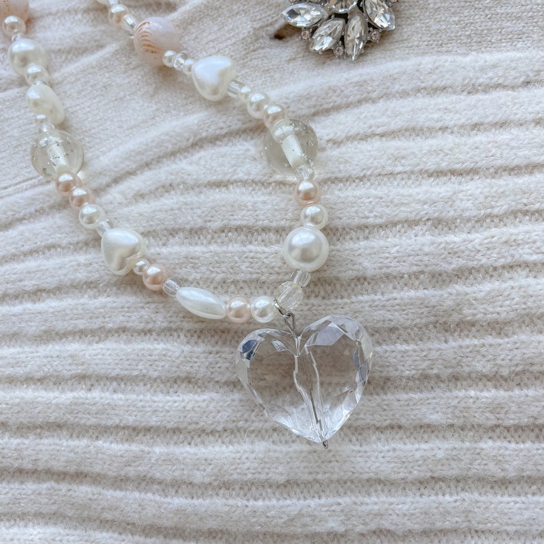 May include: A necklace with a clear, faceted heart pendant. The necklace is made with white, pink, and clear beads, including pearls and heart-shaped beads. A sparkling brooch is visible in the background.