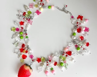 May include: A colorful necklace with a strawberry charm. The necklace is made of white, red, pink, green, and clear beads. The beads are arranged in a random pattern. The strawberry charm is red and green.