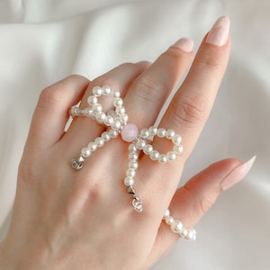 May include: A pearl ring featuring a pink gem at its centre, shaped like a bow. The ring is worn on a hand with white nail polish.