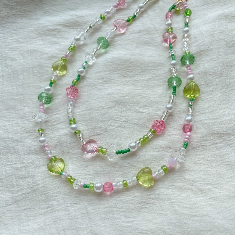 Puede incluir: Un collar de varias capas con cuentas verdes, rosas y blancas. Las cuentas tienen forma de corazones, flores y c&iacute;rculos.