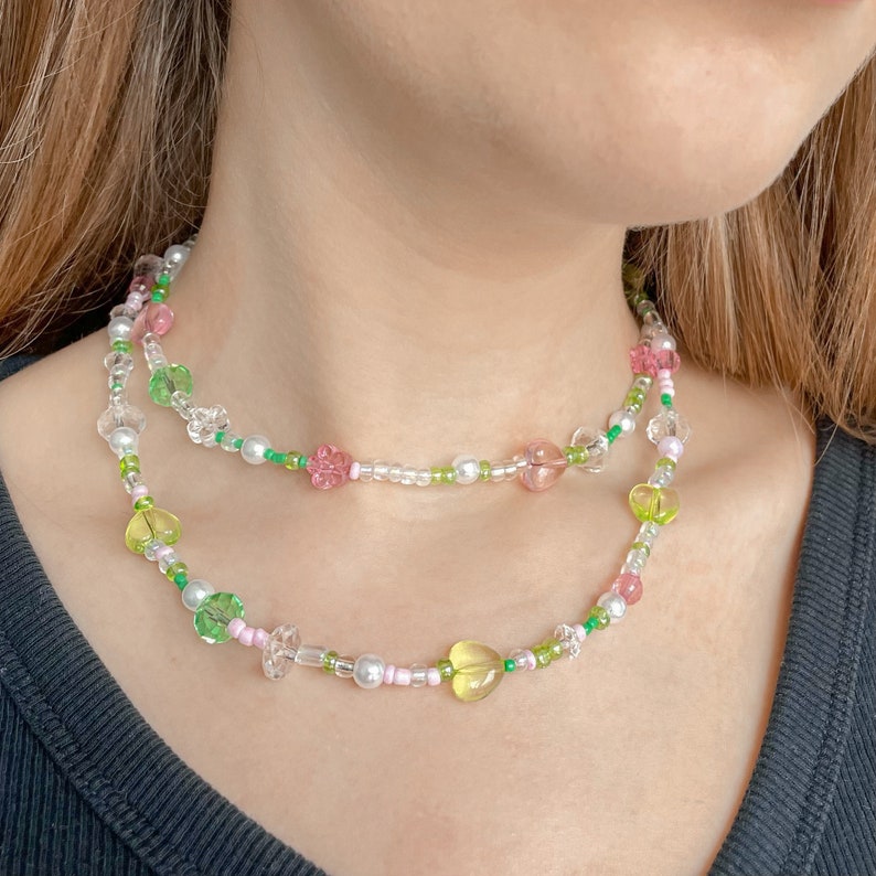Puede incluir: Un collar de dos capas con cuentas rosas, verdes y blancas. Las cuentas tienen forma de flores y corazones.