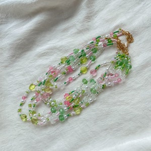 Puede incluir: Collar de m&uacute;ltiples hilos con cuentas verdes, rosas y blancas. Las cuentas est&aacute;n dispuestas en un patr&oacute;n en capas, creando un look delicado y femenino.