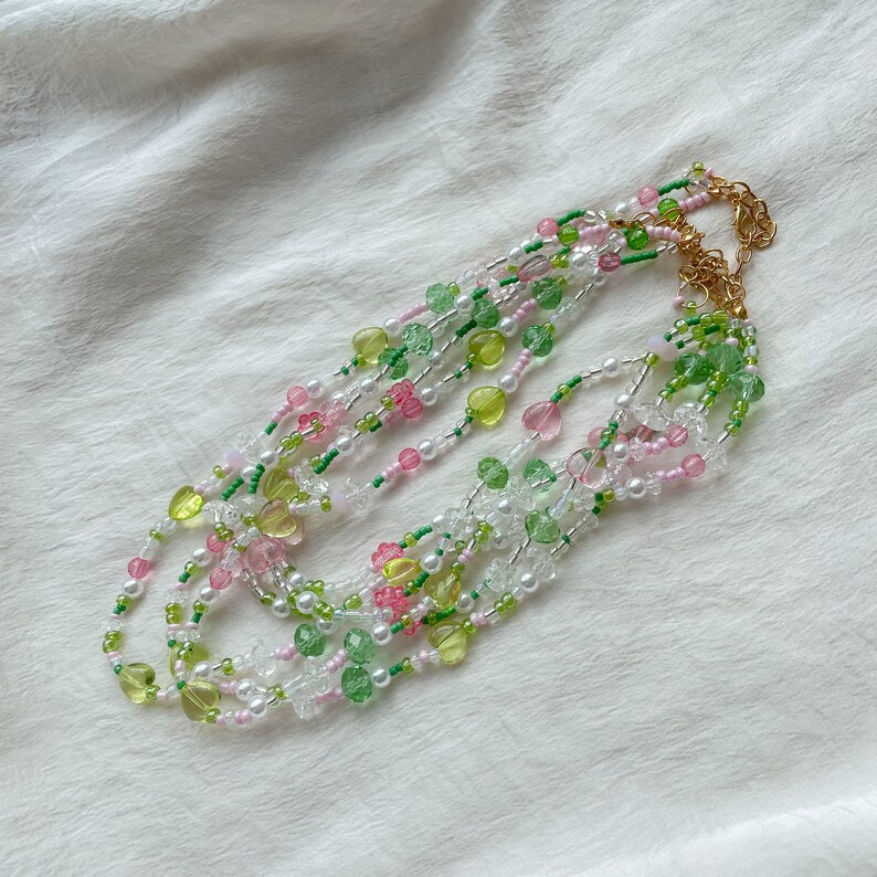 Puede incluir: Collar de m&uacute;ltiples hilos con cuentas verdes, rosas y blancas. Las cuentas est&aacute;n dispuestas en un patr&oacute;n en capas, creando un look delicado y femenino.