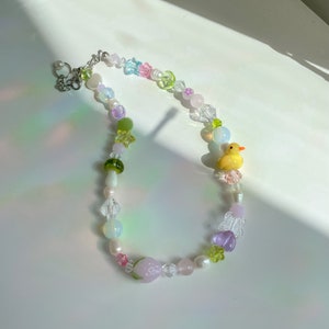 May include: A colorful beaded necklace with a variety of charms, including a yellow rubber duck, a green mushroom, a pink strawberry, and a purple butterfly. The necklace is made with clear, iridescent, and opaque beads in shades of pink, green, and blue.