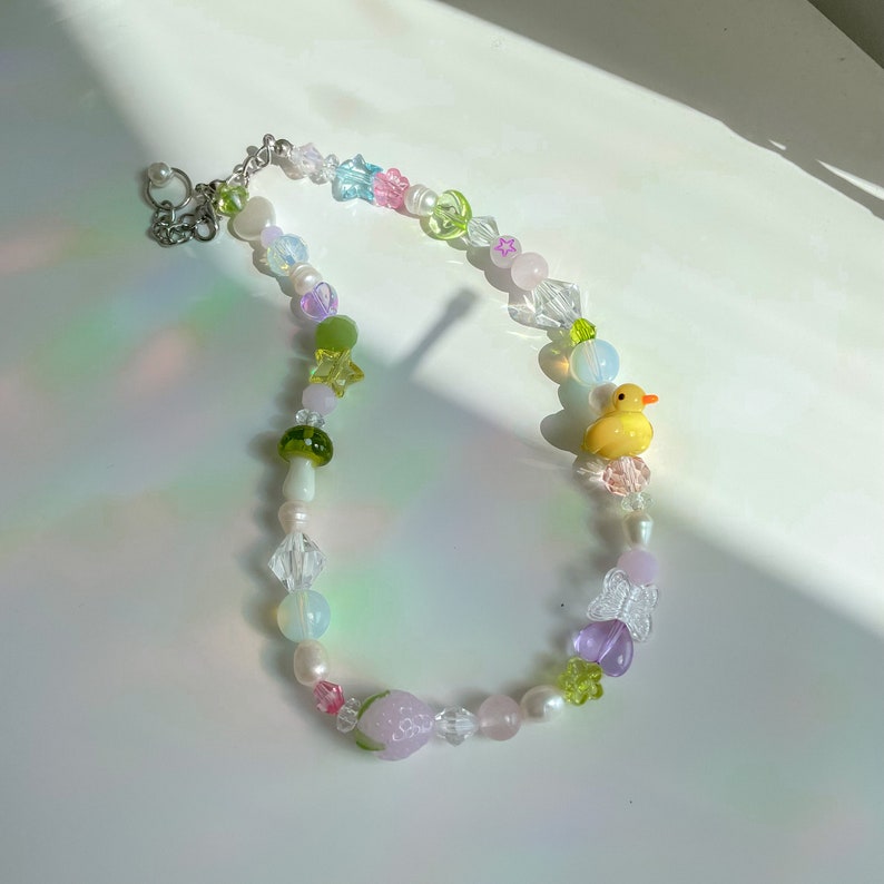 May include: A colorful beaded necklace with a variety of charms, including a yellow rubber duck, a green mushroom, a pink strawberry, and a purple butterfly. The necklace is made with clear, iridescent, and opaque beads in shades of pink, green, and blue.