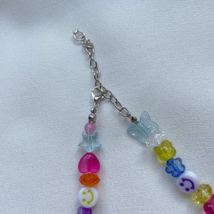 May include: A rainbow beaded necklace with a silver chain clasp. The beads are in the shape of hearts, stars, butterflies, and smiley faces. The beads are clear, pink, orange, yellow, green, blue, and purple.