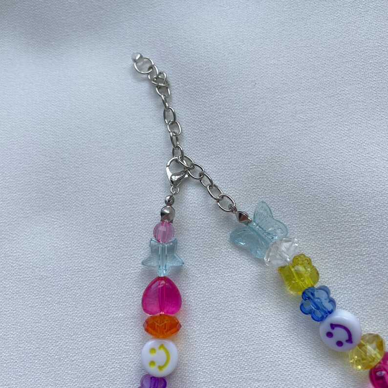 May include: A rainbow beaded necklace with a silver chain clasp. The beads are in the shape of hearts, stars, butterflies, and smiley faces. The beads are clear, pink, orange, yellow, green, blue, and purple.