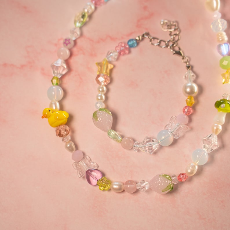 May include: A colorful beaded necklace and bracelet set with a yellow duck charm, pink and green strawberry charms, and clear crystal beads.