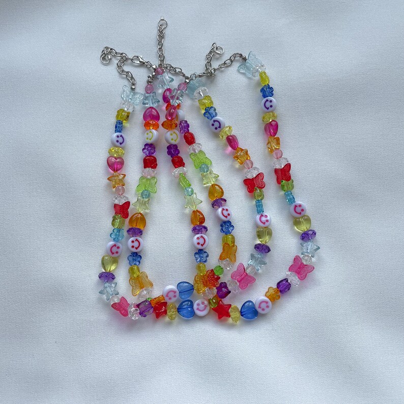 May include: Multicoloured beaded necklaces with smiley face and butterfly charms. The necklaces are made of clear plastic beads with rainbow colours and silver metal clasps.
