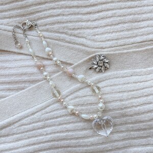 May include: A necklace with a clear heart pendant, white and pink beads, and a silver clasp. A silver ring with a flower design is also present. The jewellery is displayed on a cream-coloured, ribbed fabric.