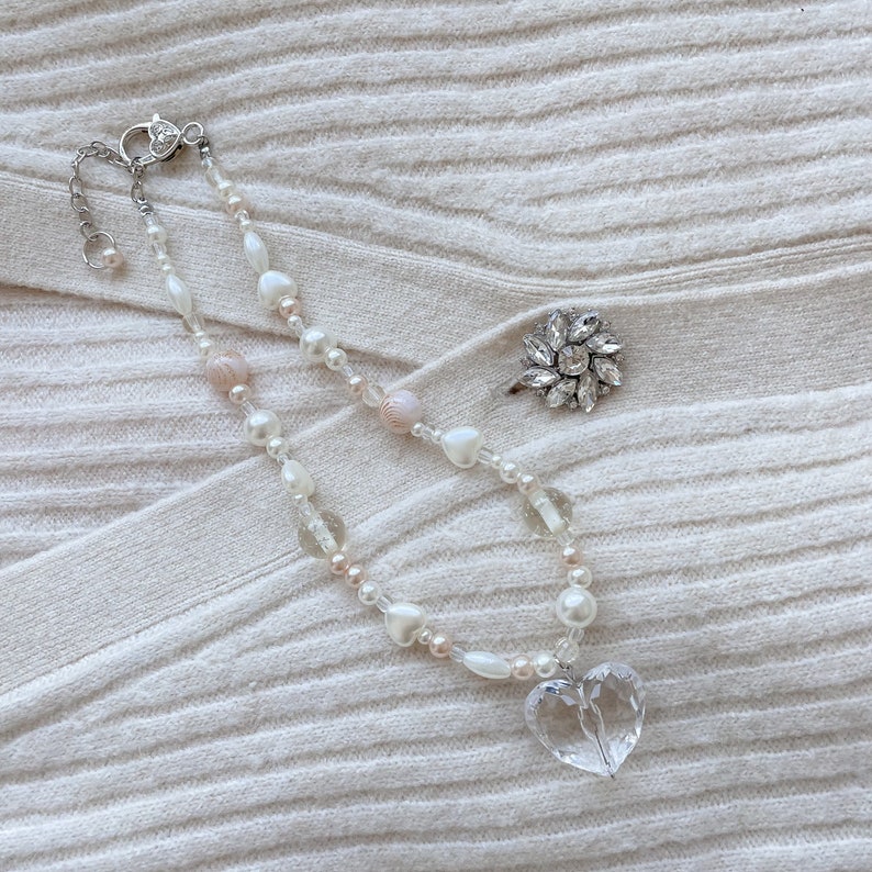 May include: A necklace with a clear heart pendant, white and pink beads, and a silver clasp. A silver ring with a flower design is also present. The jewellery is displayed on a cream-coloured, ribbed fabric.