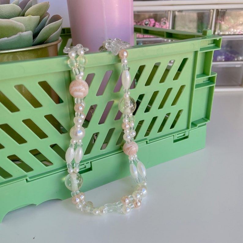 Peut inclure: Un collier de perles blanches et roses avec des perles de verre transparentes et une caisse de rangement en plastique verte en arri&egrave;re-plan.