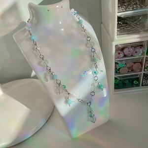 Pode incluir: Um delicado colar de corrente prateada com pingentes e contas de borboleta azul claro e transparente. O colar tem um pingente de cristal.