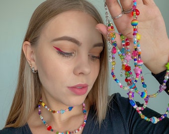 Op de afbeelding: Een close-up van een vrouw die een zwart shirt draagt en een kleurrijke kralenketting vasthoudt. De ketting is gemaakt van verschillende kralen, waaronder sterren, harten en regenboogkleurige kralen.