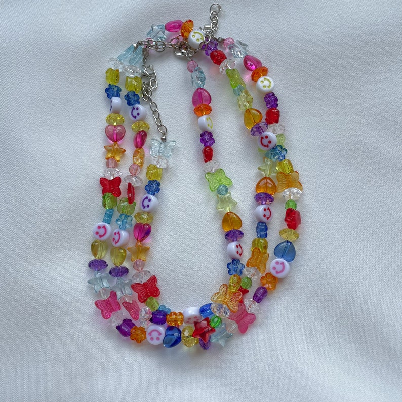 May include: Multi-layered necklace featuring colourful beads, including butterflies, stars, hearts, and smiley faces. The necklace is made of plastic and has a silver chain.