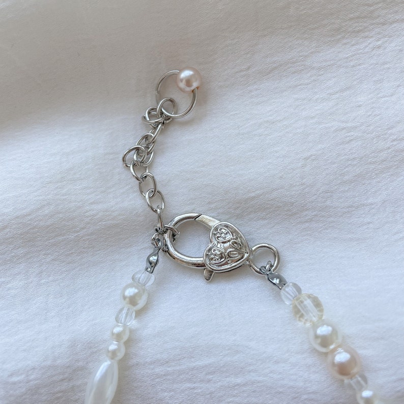 May include: Close-up of a pearl necklace with a silver chain and clasp. The necklace features a variety of white and off-white pearls and beads. The clasp is heart-shaped.