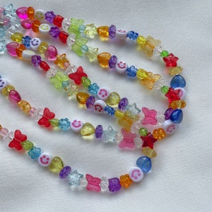 May include: A colourful beaded necklace featuring smiley face charms, butterflies, stars and hearts. The necklace is made from a variety of colours, including red, orange, yellow, green, blue and purple.