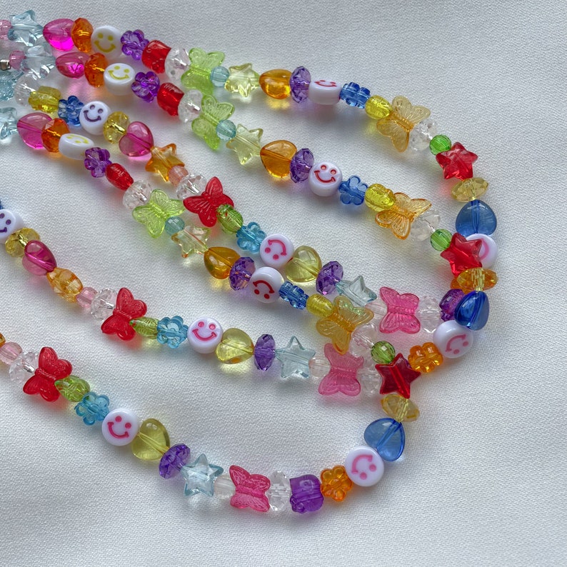 May include: A colourful beaded necklace featuring smiley face charms, butterflies, stars and hearts. The necklace is made from a variety of colours, including red, orange, yellow, green, blue and purple.