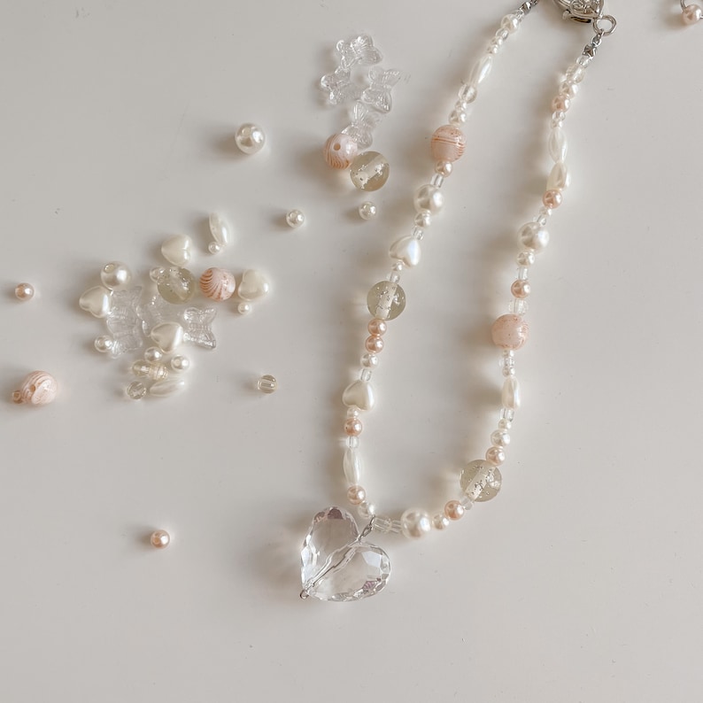 May include: A necklace with a clear heart pendant, strung with a mix of white, pink, and clear beads. The design includes pearls and heart-shaped beads. The clasp is silver. Additional beads of different shapes and sizes are scattered around the necklace.