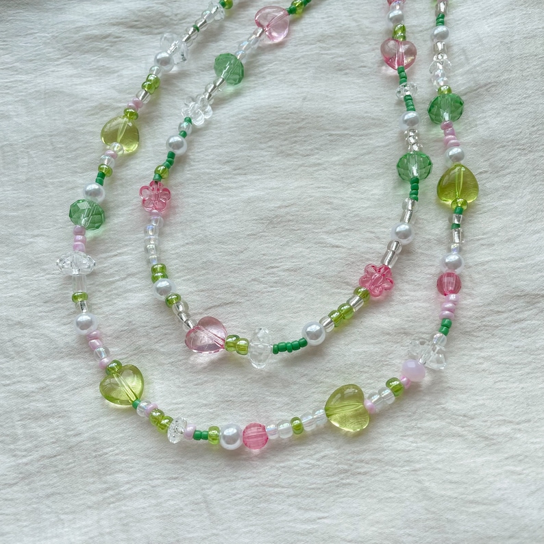 Puede incluir: Dos collares superpuestos con cuentas verdes, rosas y blancas. Los collares presentan cuentas en forma de coraz&oacute;n y flor.
