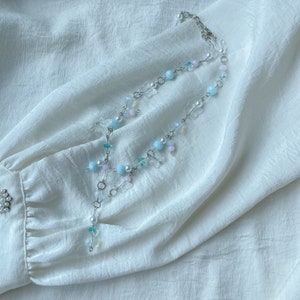 Pode incluir: Um delicado colar de corrente prateada com pingentes e contas de borboleta em azul pastel, rosa e branco. O colar est&aacute; sobre um fundo de tecido branco.