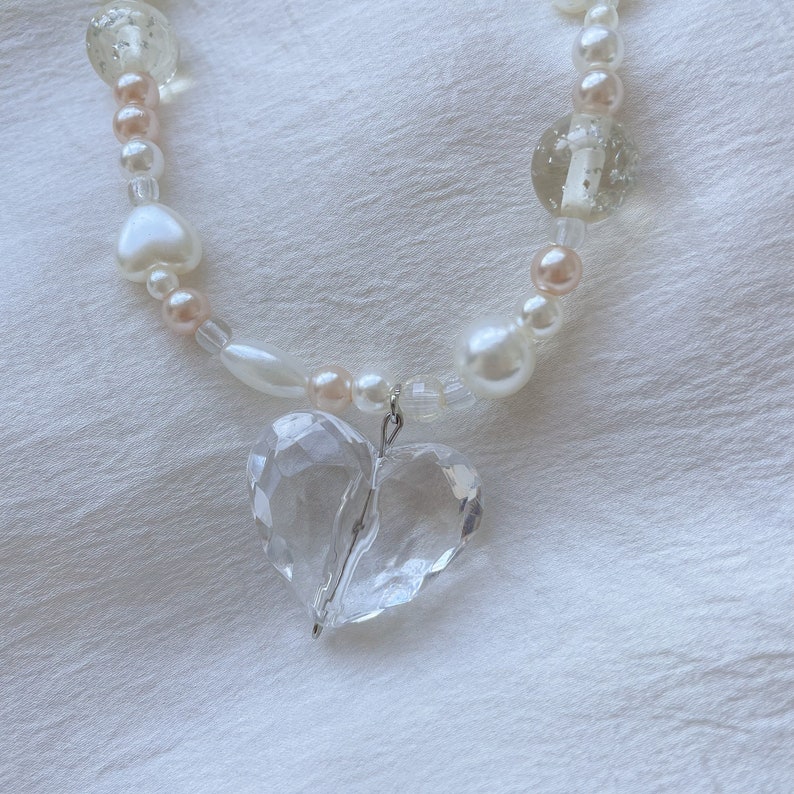 May include: A necklace with a clear, faceted heart pendant. The necklace features white, peach, and clear beads, including pearls and heart-shaped beads. The necklace is displayed on a white, textured surface.