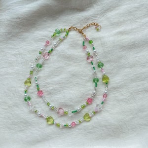 Puede incluir: Un delicado collar de dos capas con cuentas de coraz&oacute;n de color rosa, verde y blanco. El collar est&aacute; hecho de cuentas de vidrio y tiene un cierre dorado.