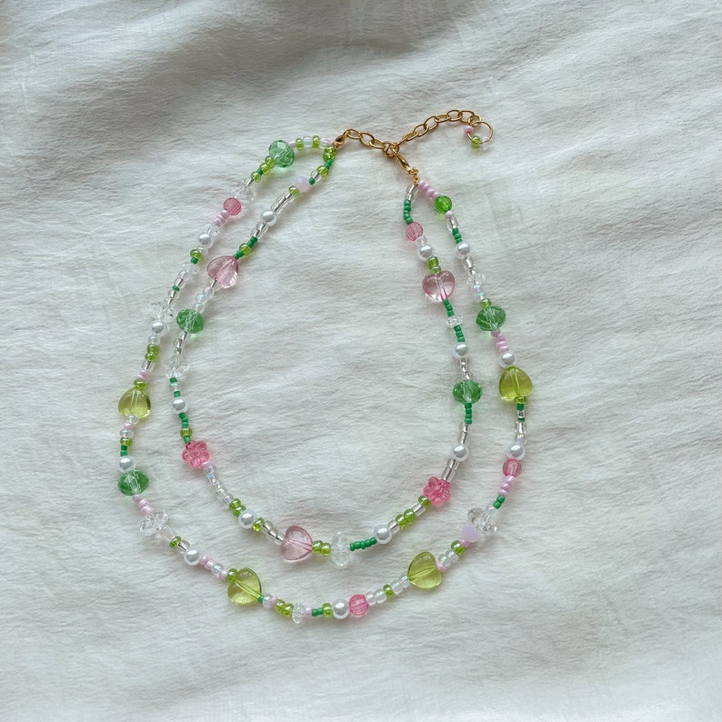 Puede incluir: Un delicado collar de dos capas con cuentas de coraz&oacute;n de color rosa, verde y blanco. El collar est&aacute; hecho de cuentas de vidrio y tiene un cierre dorado.