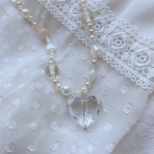 May include: A necklace with a clear, heart-shaped crystal pendant. The chain is made of white pearls, pink beads, and clear glass beads. The pendant hangs from a silver-coloured bail, creating a romantic aesthetic.