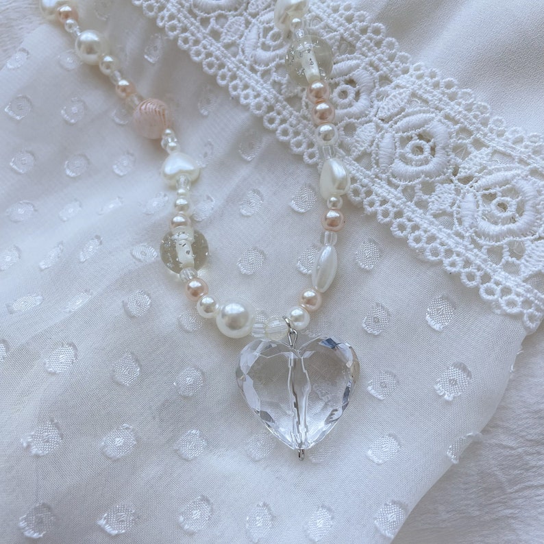 May include: A necklace with a clear, heart-shaped crystal pendant. The chain is made of white pearls, pink beads, and clear glass beads. The pendant hangs from a silver-coloured bail, creating a romantic aesthetic.