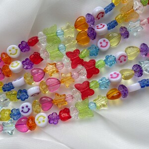 May include: A colourful beaded necklace featuring a variety of shapes, including hearts, stars, butterflies, and smiley faces. The beads are made from clear plastic and are strung on a clear elastic cord.