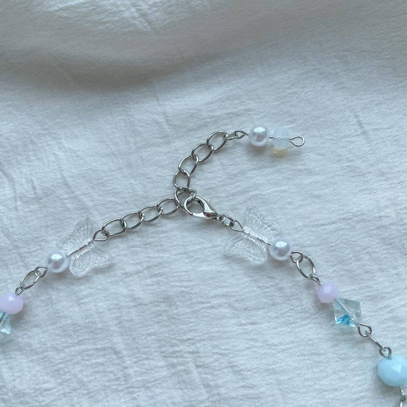 Pode incluir: Um delicado colar de corrente prateada com fecho de lagosta. O colar apresenta pingentes de borboleta de pl&aacute;stico transparente, contas de p&eacute;rola branca e contas de cristal azul claro e rosa.