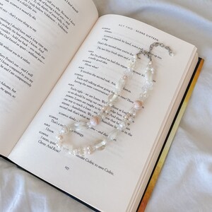 Peut inclure: Un collier de perles blanches et roses avec des coquillages et des perles de verre transparentes. Le collier est pos&eacute; sur un livre ouvert avec du texte sur une pi&egrave;ce de th&eacute;&acirc;tre. Le texte du livre indique "Acte Deux Sc&egrave;ne Seize".