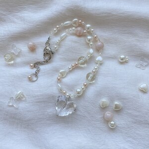 May include: A delicate necklace with a clear heart pendant and a strand of white and pink beads. The necklace includes clear glass beads, small white pearls, and heart-shaped beads. Clear butterfly and heart-shaped embellishments are scattered around.