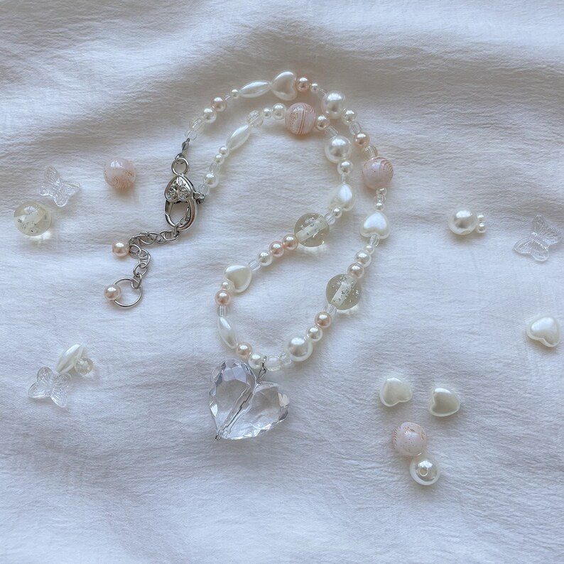 May include: A delicate necklace with a clear heart pendant and a strand of white and pink beads. The necklace includes clear glass beads, small white pearls, and heart-shaped beads. Clear butterfly and heart-shaped embellishments are scattered around.
