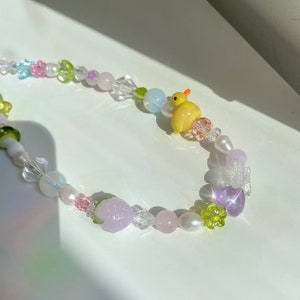 May include: A colorful beaded necklace with a variety of shapes and sizes, including a small yellow duck charm. The necklace features pastel pink, green, blue, and white beads.