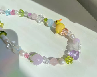 May include: A colorful beaded necklace with a variety of shapes and sizes, including a small yellow duck charm. The necklace features pastel pink, green, blue, and white beads.