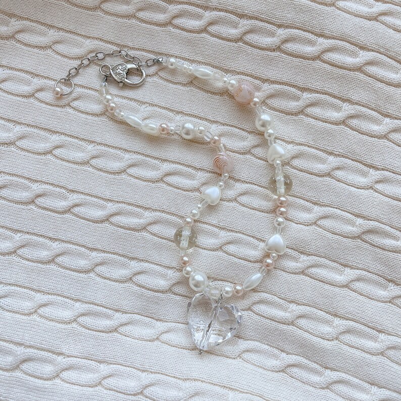 May include: A necklace with a clear heart pendant, strung with white pearls, clear beads, and small pink beads. The silver-toned clasp secures the necklace. The jewellery is displayed on a white, textured background.